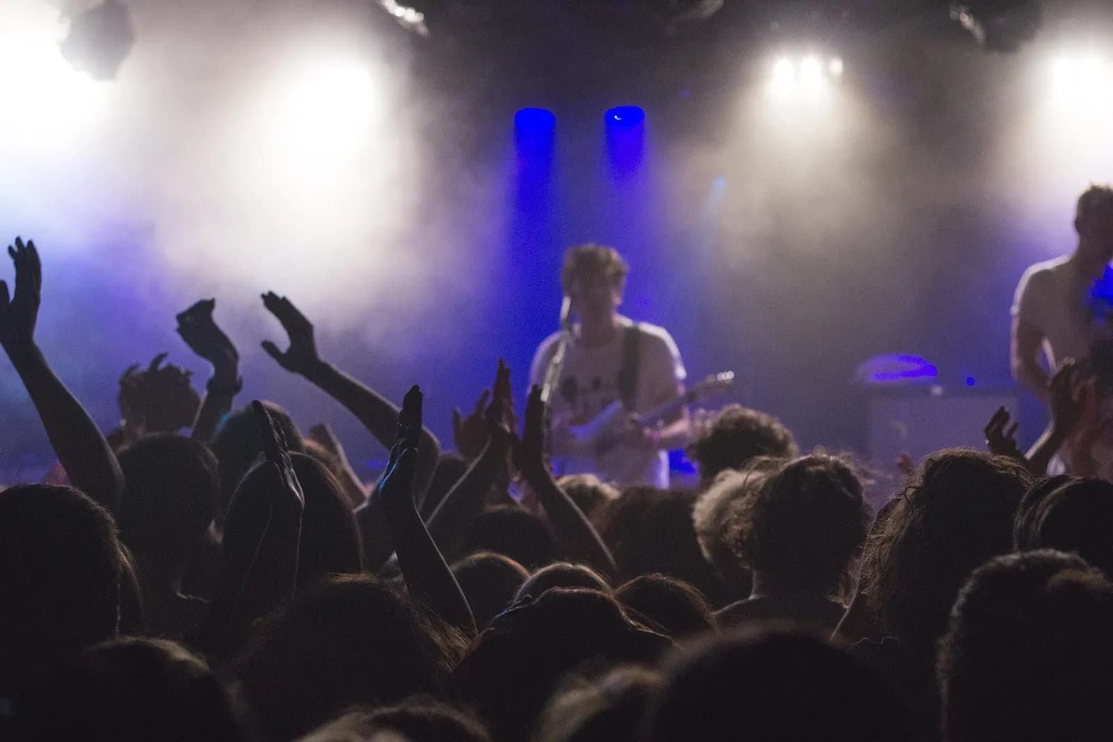 Excited audience with raised hands in front of a stage lit by blue and white spotlights